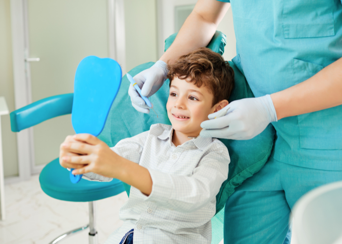 Child in dental chair holding blue mirror, smiling at dental professional in scrubs, emphasizing pediatric dental care and positive experiences in children's dentistry.