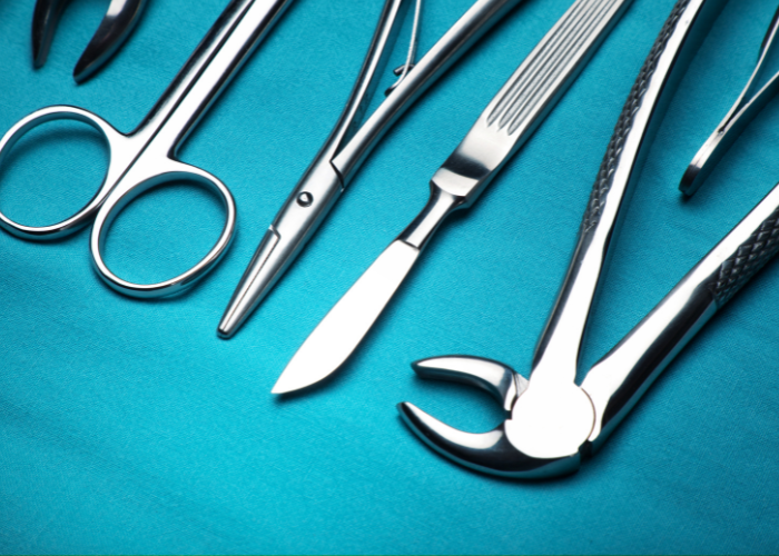 Dental surgical instruments on a teal surface, including forceps, scissors, a scalpel, and other tools used for oral surgery and extractions.