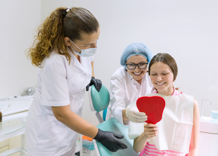 Teen receiving dental care, smiling while holding a heart-shaped mirror, with two dental professionals assisting, in a modern dental office setting.